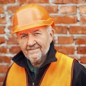Senior male foreman in hardhat standing at masonry brick wall at building new house. Portrait of happy man engineer or construction worker at construction site